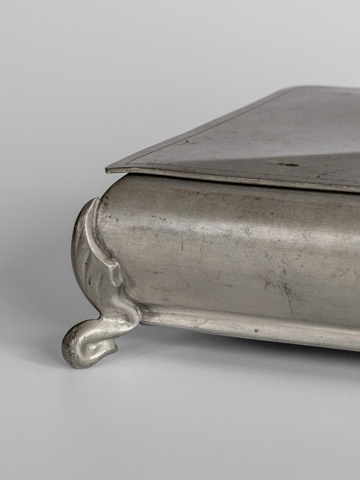 A close-up of the Spigel Art Deco Metal Box reveals its vintage silver allure, complete with an ornate foot and a subtly tarnished surface against a neutral backdrop. The box features a partially open hinged lid, highlighting its intricate design details reminiscent of the Art Deco style.