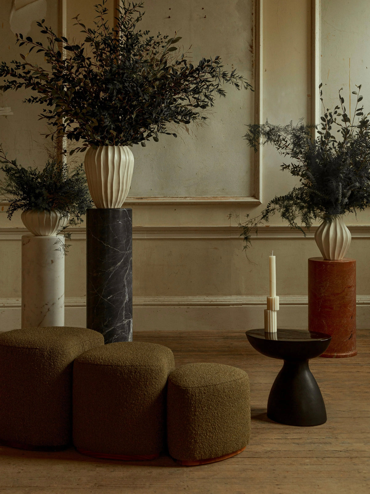 A stylish room showcases three tall vases in different colors filled with lush greenery. In the foreground, a set of rounded, olive-colored seating encircles an asymmetric matte black table adorned with tall candles, all set against a textured wall. The "Cleo" Ceramic Side Table by Ransom & Dunn adds charm to the scene.