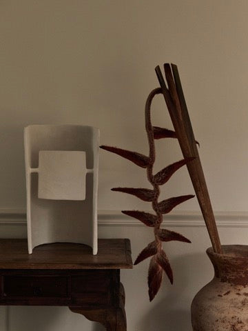 A wooden table with a white chair sculpture is softly illuminated by Studio Tristan Kallas' Crescent Table Lamp, casting a gentle glow on a vase with a dried red flower and two wooden sticks. The cream-colored wall backdrop enhances the serene and rustic ambiance.