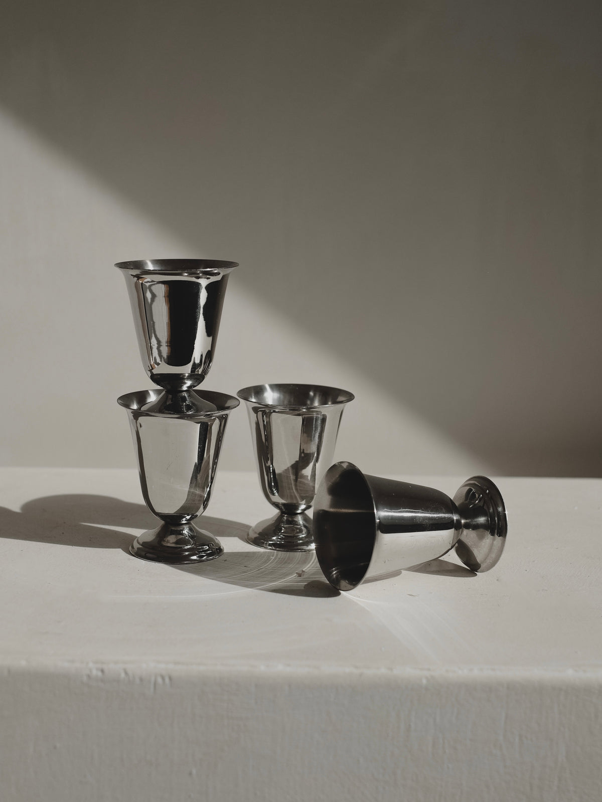 A Set of 4 Coupes by Guy Degrenne from septembre studios is displayed on a white surface: three are upright and stacked, the fourth lies on its side. Soft lighting emphasizes their classic 1970s stainless steel design.
