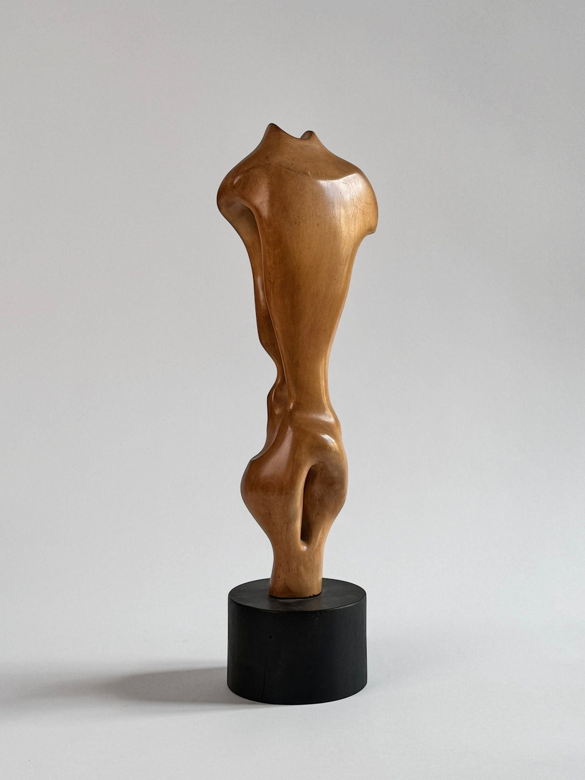 The Figuresdesign "Wood Sculpture by Pierre Dedieu" features a smooth, abstract design with an organic shape reminiscent of a torso. This unique piece, crafted from varnished walnut wood, showcases flowing curves and a polished finish. It stands gracefully on a rounded black base against a neutral gradient background.