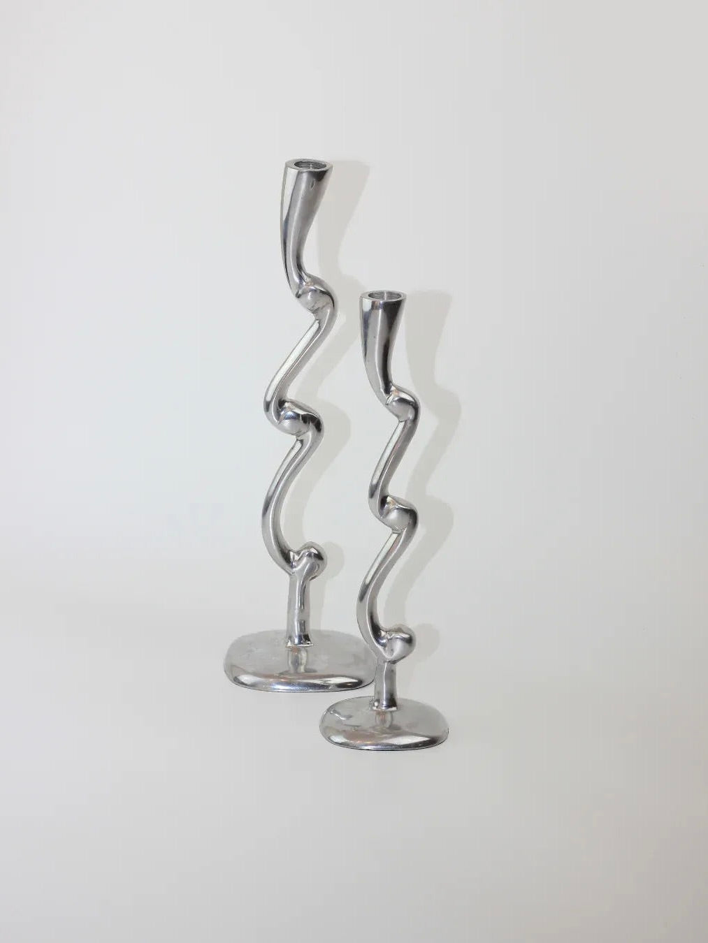 A pair of aluminum candleholders from Boga Avante Shop, featuring wavy, abstract designs, stand on a light gray surface against a white background, bringing a modern touch to any setting.