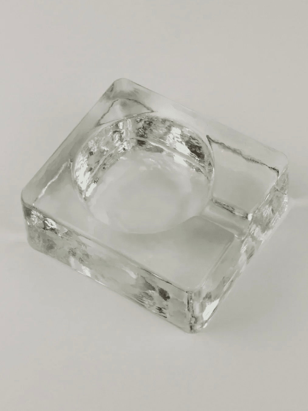 The Brutalist Ashtray by Boga Avante Shop is a clear, rectangular glass piece featuring a round indentation for holding ashes and a small groove on one side for resting a cigarette. Exhibiting hints of Brutalist style, this vintage ashtray makes an elegant yet understated statement when placed on a white surface.