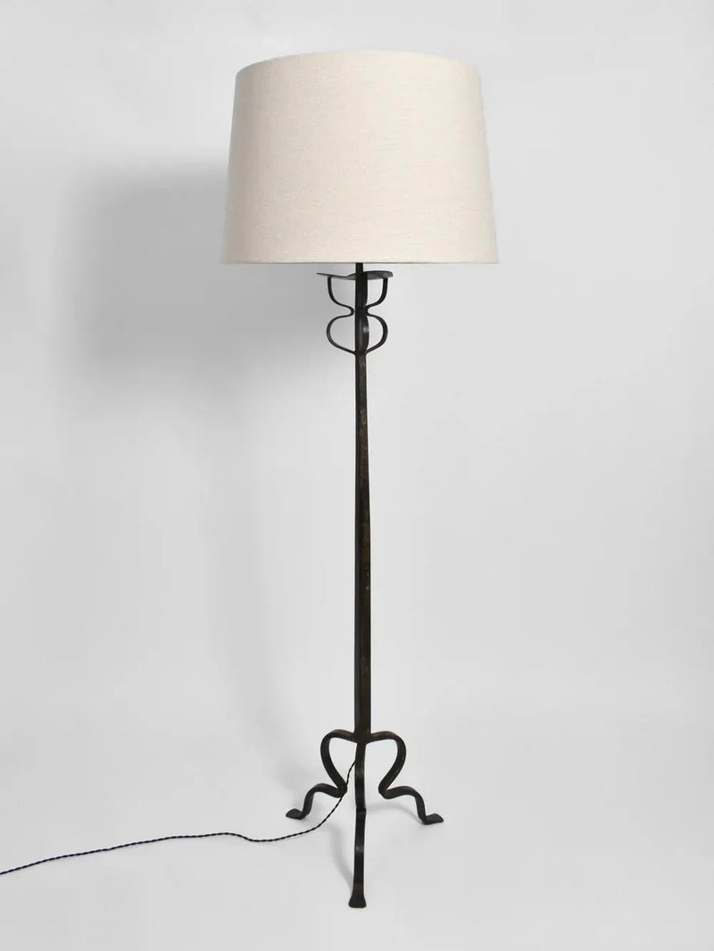 The Tripod Iron Floor Lamp from Barracuda Interiors features a tall, slender stand with a graceful curved design at the base, crafted in wrought iron. It is topped with a beige cylindrical lampshade and has an elegantly extending black power cord, reminiscent of 1960s France.