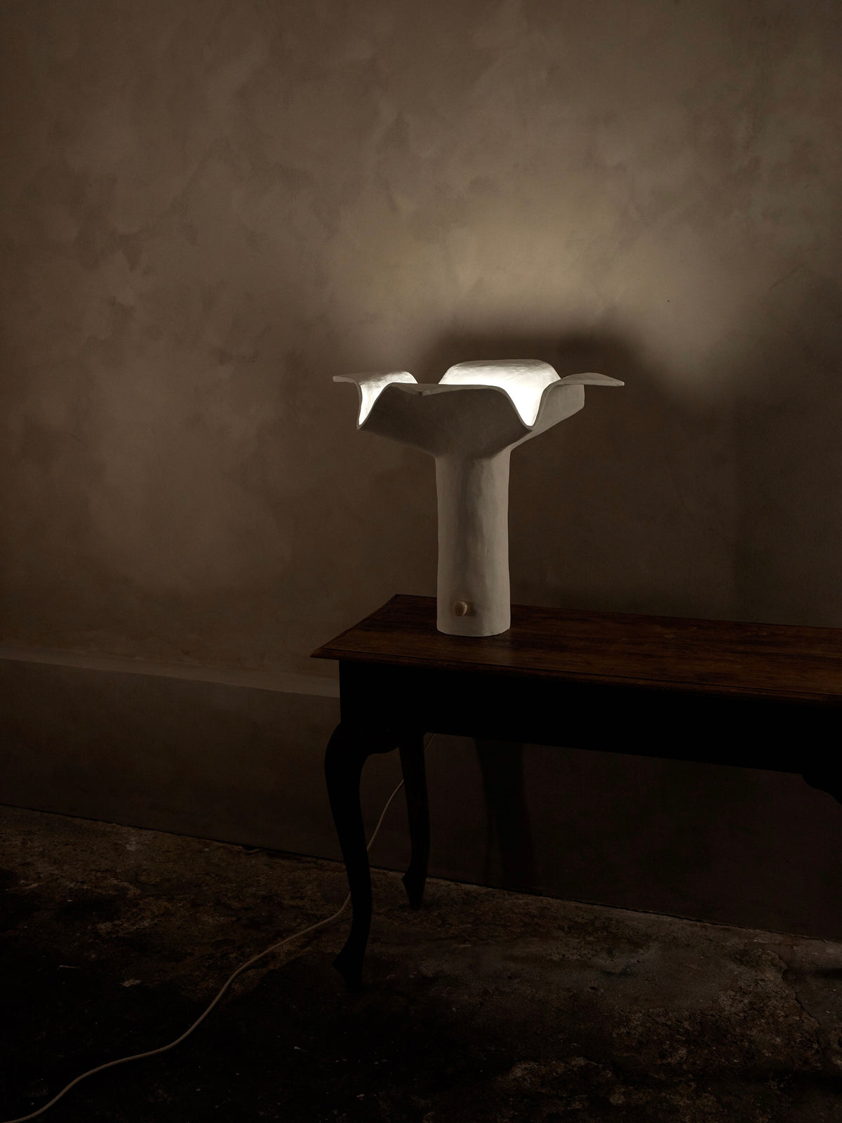 The La Fleur Table Lamp by Studio Tristan Kallas casts a soft glow on a wooden table against a textured, dimly lit wall. Its artistic form features floral silhouettes with a broad, flat top and slender base. A visible power cord adds to its sculptural design.
