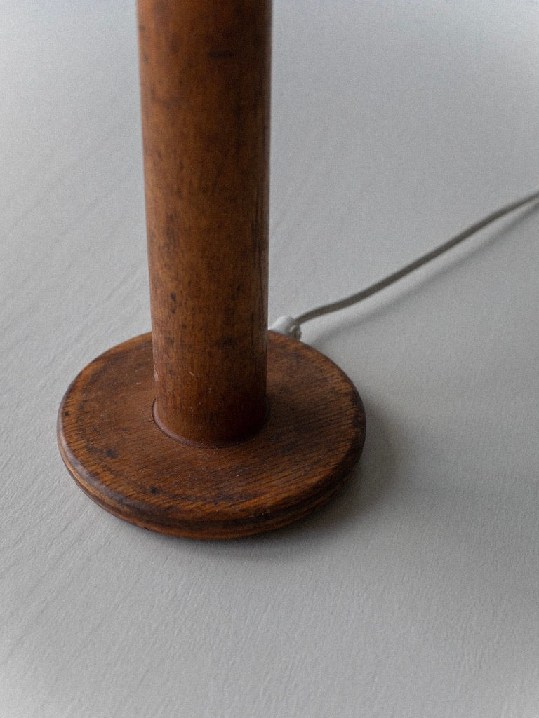 A close-up of the base of the Midcentury Table Lamp by Out For Lunch, showcasing a cylindrical wooden post affixed to a round wooden platform. The cord extends from the wooden handle base, which is partially visible on a light-colored surface.