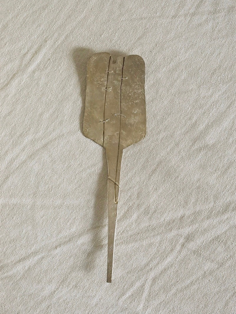 The Silver Plated Cake Server by Rooms Studio, featuring a long handle and a rectangular serving area reminiscent of a vintage design, rests elegantly on a light beige, textured fabric background. The serving area is securely attached to the handle, exuding an old-fashioned charm.