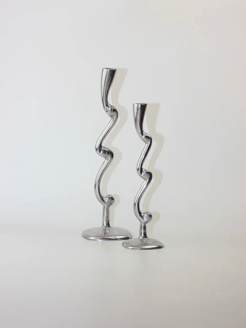 A pair of aluminum candleholders from Boga Avante Shop features curvy, abstract designs and stands against a plain white background. The unique wavy shapes of these candleholders add a modern, sophisticated touch, creating an artistic and sculptural design.