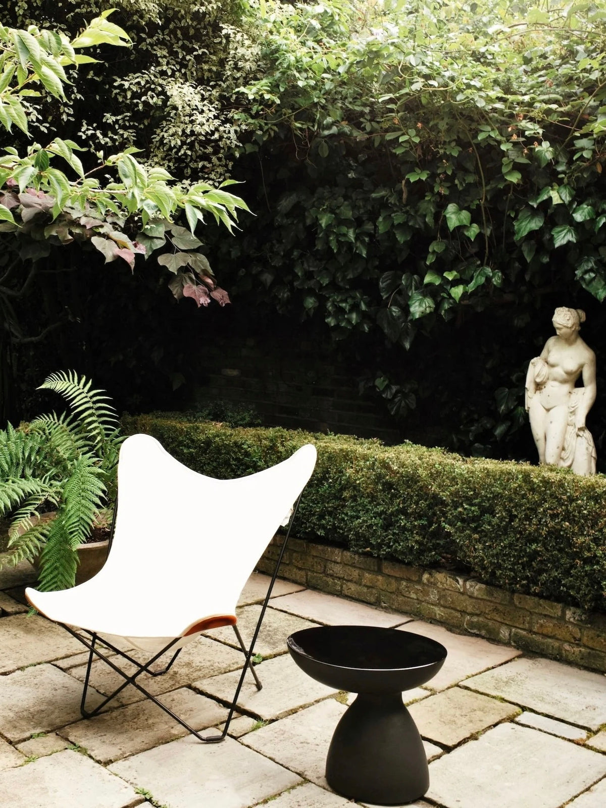 A garden setting features the "Cleo" Ceramic Side Table by Ransom & Dunn alongside a white butterfly chair on a stone patio. The area is enveloped in lush greenery, including ivy and ferns, while a classic statue peeks through dense foliage and neatly trimmed hedges.