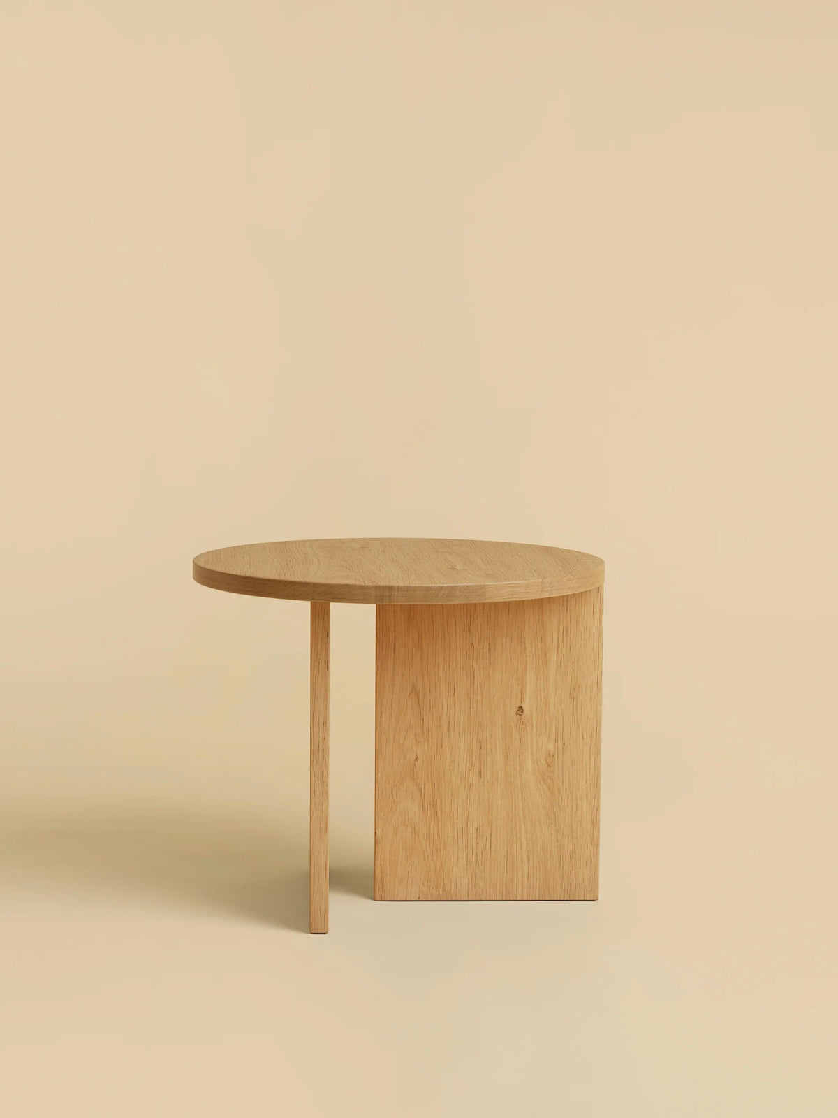 The "Kume" Side Table by Lebanto features a minimalist design with a semi-circular solid oak top and two legs, one flat and one slender. Set against a beige background, it highlights the natural wood texture and simplicity.