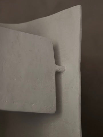 A minimalist abstract sculpture with interlocking rectangles and a peg detail resembles the Crescent Table Lamp by Studio Tristan Kallas. Its smooth, monochromatic gray surface creates an effect similar to indirect lighting.