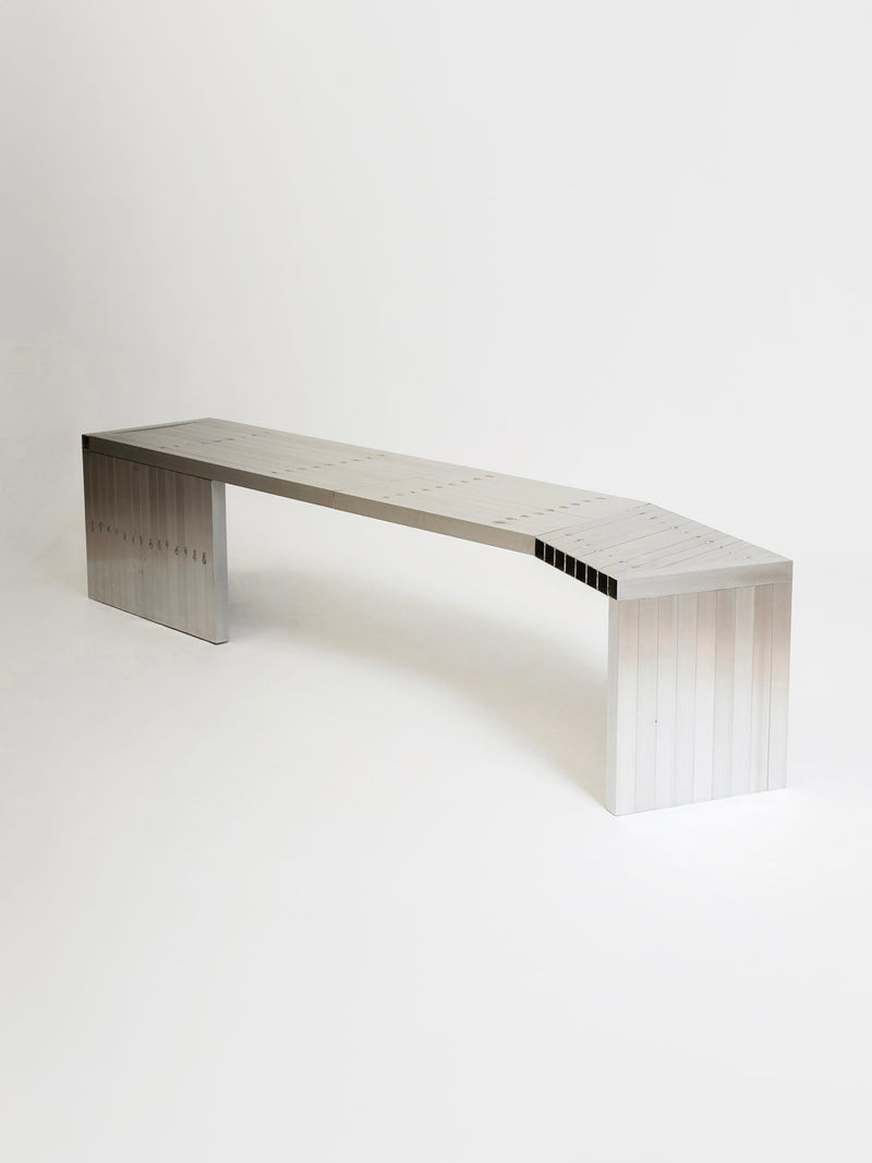The Aluminum Bench by Studio HAOS features a minimalist, modern design with clean lines, a rectangular seat with subtle grooves, and two flat vertical legs, all set against a plain white background.