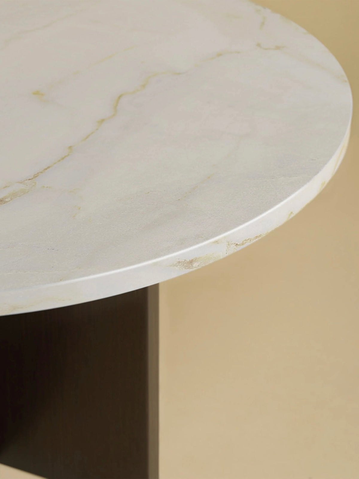 A close-up of the "Tinian" Coffee Table by Lebanto showcases a round white marble top with subtle gray and gold veining, crafted through artisan production. The dark oak wood base is visible against a beige background.