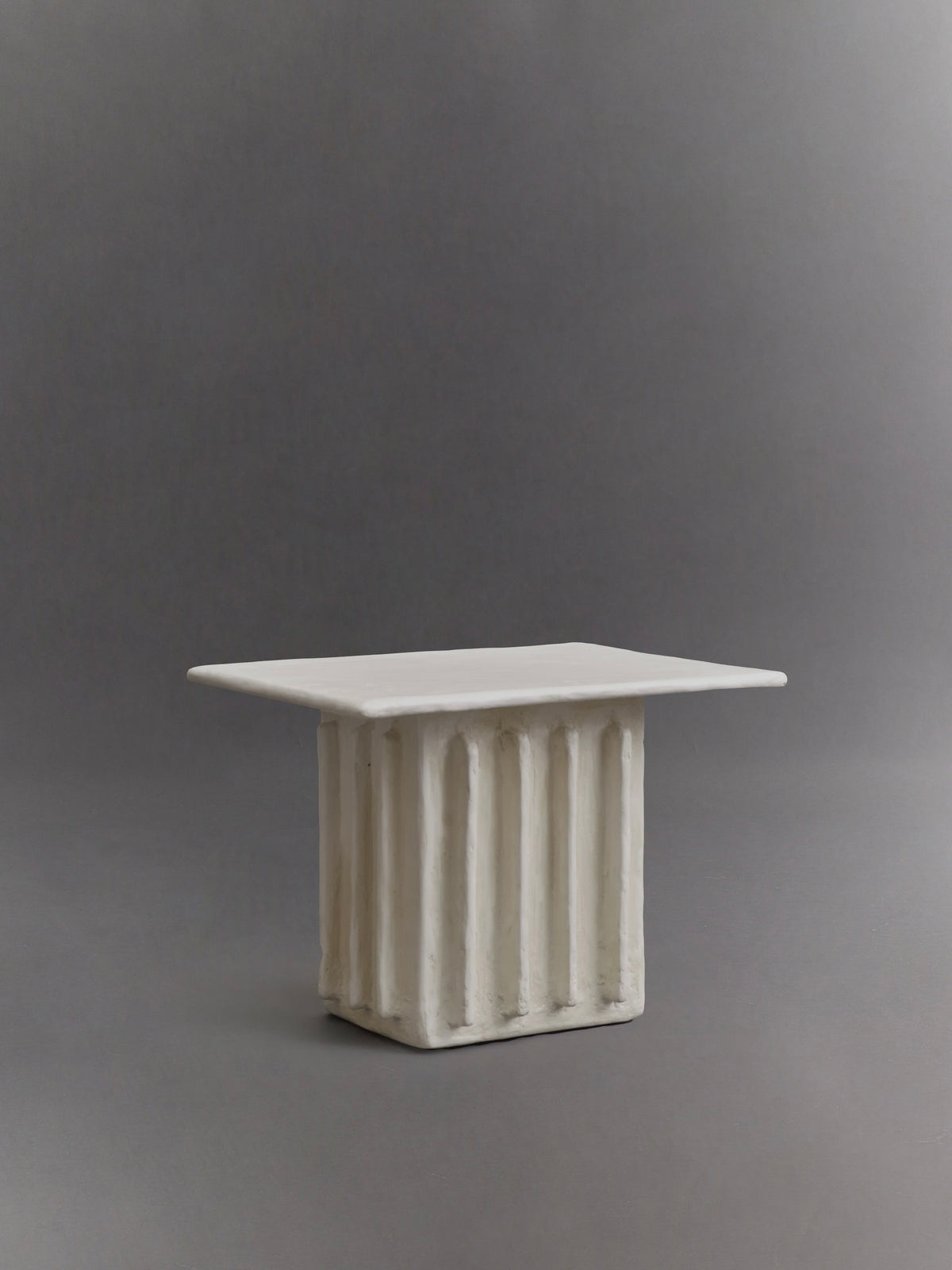 The Columnar Side Table by Ombia Studio is a rectangular, sculpted piece made from raw white plaster with vertical grooves, resembling a small table or pedestal, showcased against a neutral gray background.