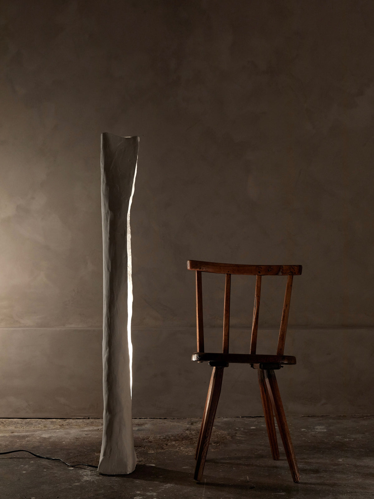 A wooden chair sits beside the Alcove Floor Lamp, its soft light enhancing a minimalist setting. The lamp's organic cove shape complements muted tones and textured walls, reflecting Studio Tristan Kallas' refined aesthetic.