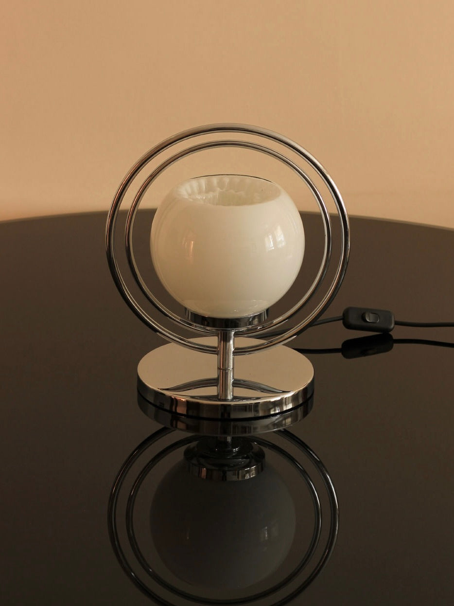 A Boga Avante Shop Space Age Table Lamp, featuring a spherical glass shade encased in concentric metal rings, rests on a glossy dark surface. The lamp has a metallic base and the black power cord with its switch is visible beside it. The reflection of the lamp on the surface below enhances its mid-century modern charm.