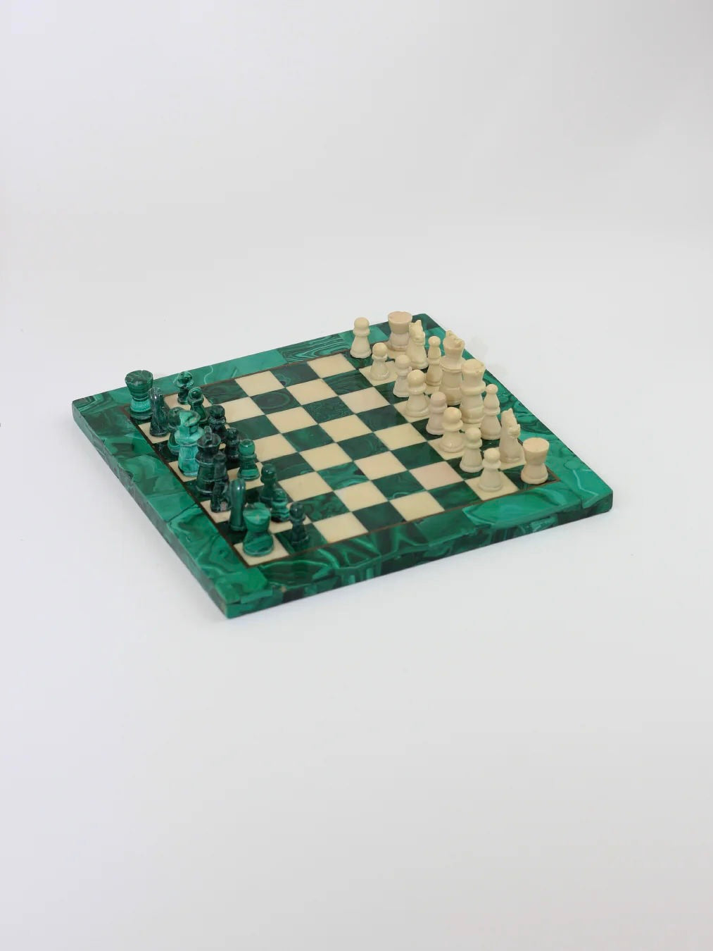 The Boga Avante Shop's Malachite And Marble Chess set showcases a remarkable green and white marble board, complete with intricately crafted green pieces on the left side and white ones on the right, perfectly arranged for an engaging match. The board is angled to emphasize its unique color palette and sophisticated design.