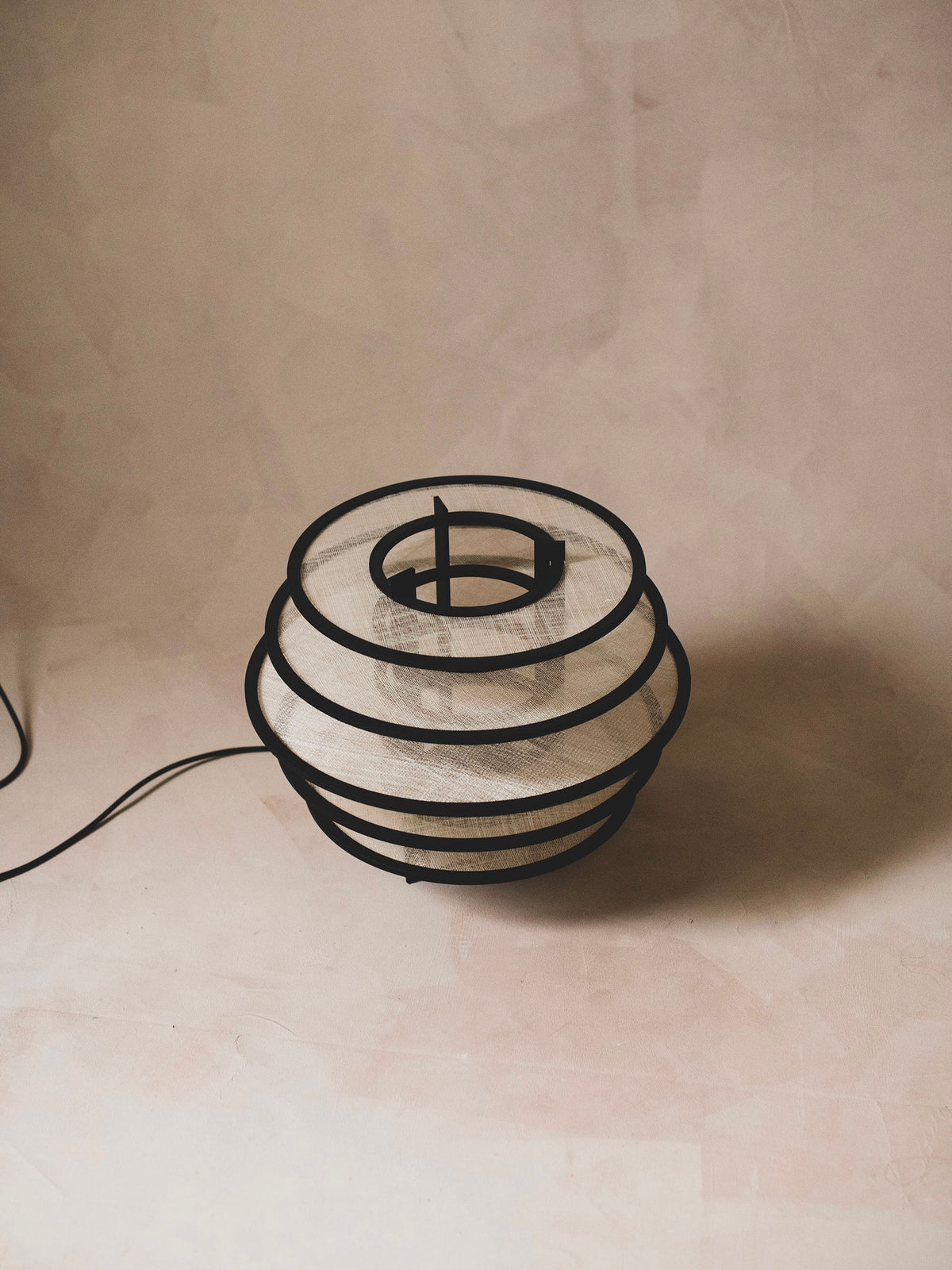 The Lamp I by ELIURPI showcases a sculptural, minimalist design with concentric black metal rings and a textured white shade, providing ambient light against a soft, neutral background.