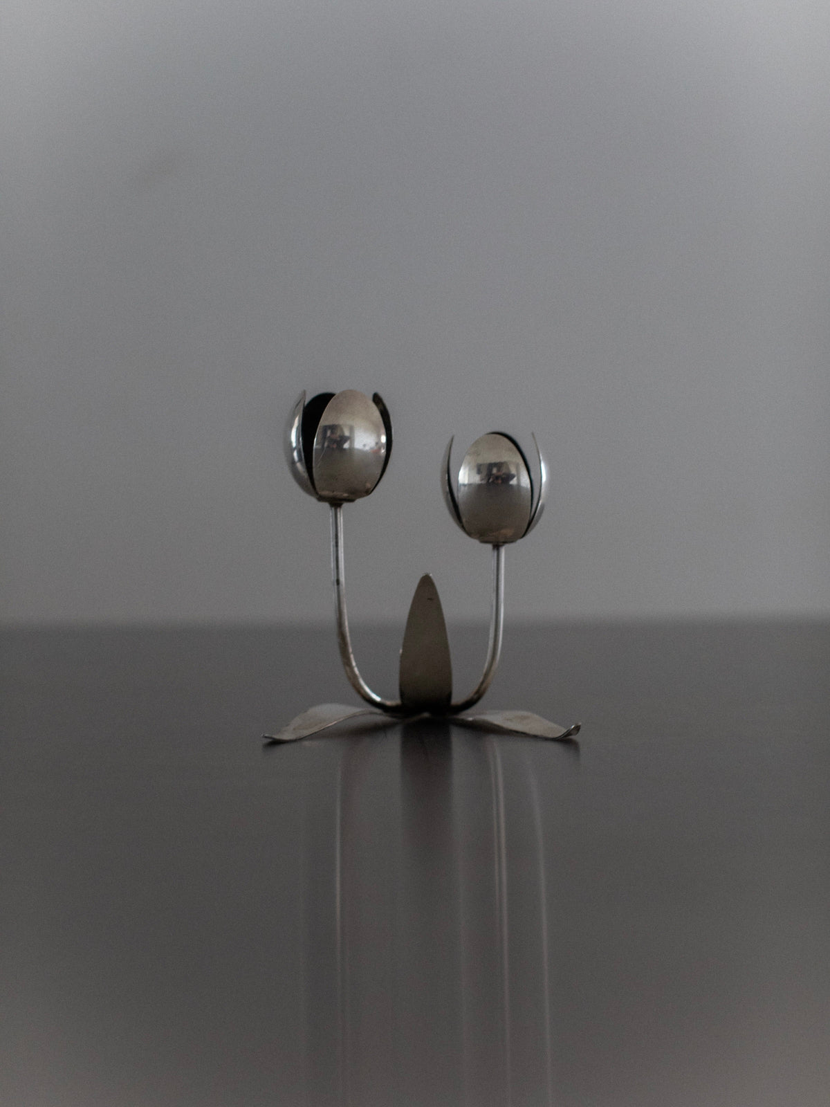 The Steel Tulip Candleholder by Out For Lunch is a captivating piece of modern home decor. This unique metal sculpture, resembling two spoon-like flowers, showcases rounded forms akin to petals or seed pods in a sleek steel design. It features a central stem with a sturdy base set against a soft gray background.