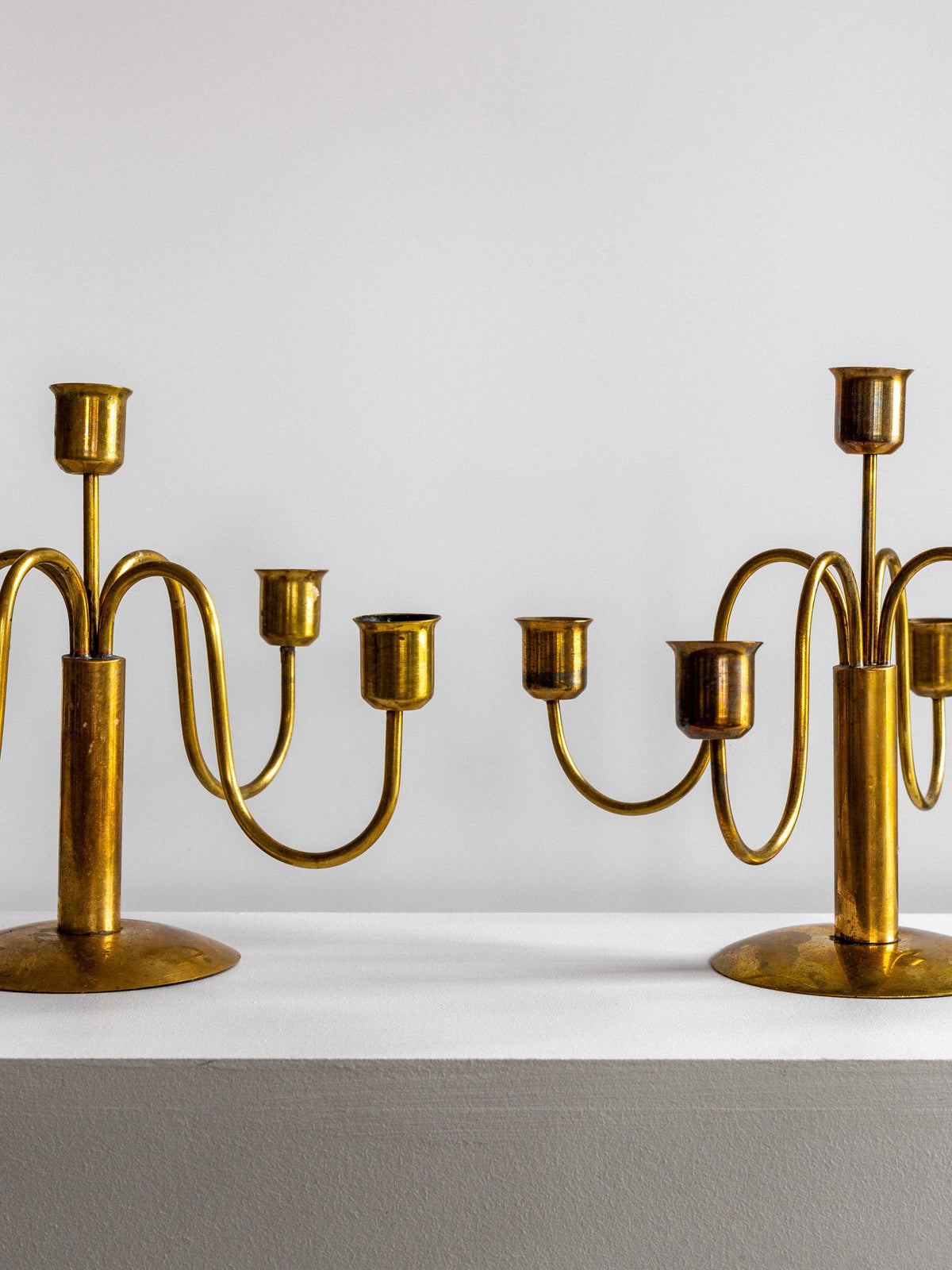 A pair of Spigel Mid-Century Brass Candelabras with a subtle patina are displayed on a minimalist white shelf against a plain white wall. Each candelabra has three arms and an ornamental design, creating a symmetrical and elegant look.