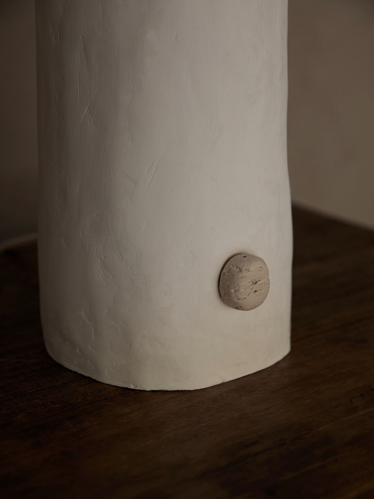 A close-up of a ceramic white cylindrical object, reminiscent of the 'La Fleur' Table Lamp by Studio Tristan Kallas, with a small round side knob. It features a rough, textured finish and rests on a dark wooden surface.