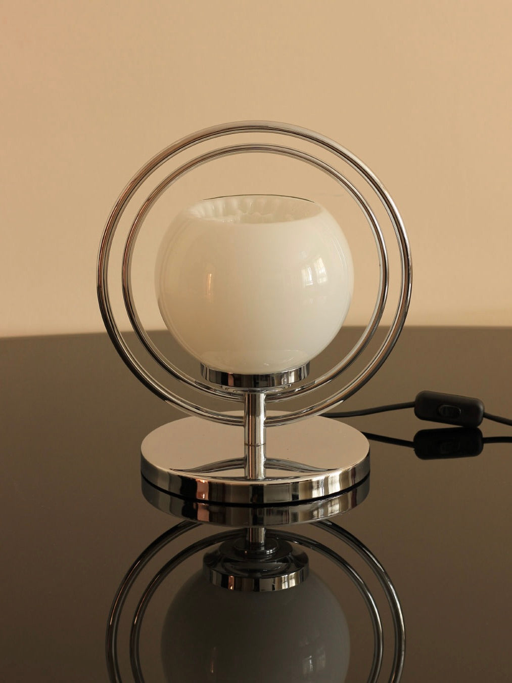 Introducing the Boga Avante Shop's Space Age Table Lamp—a sleek, modern design with a chrome circular stand and a white frosted spherical glass shade. This stylish lamp rests on a reflective surface and features a black power cord with an inline switch for easy operation.