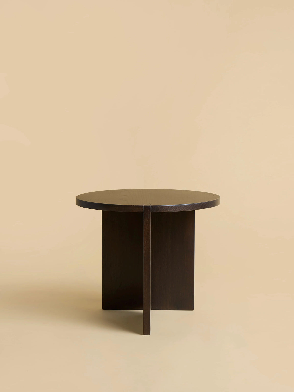 The "Kume" Side Table by Lebanto features a minimalist round design with a solid oak wood top and dark finish, complemented by a modern cross-shaped base. Its neutral beige background highlights the simplicity and elegance of this sophisticated piece.