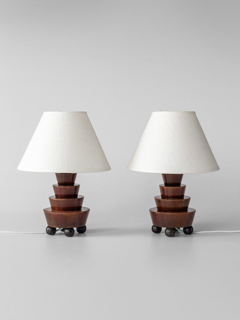 Two Spigel Art Deco Wooden Table Lamps with tiered geometric brown bases and white fabric cone shades are plugged in and displayed side by side against a plain light gray background.
