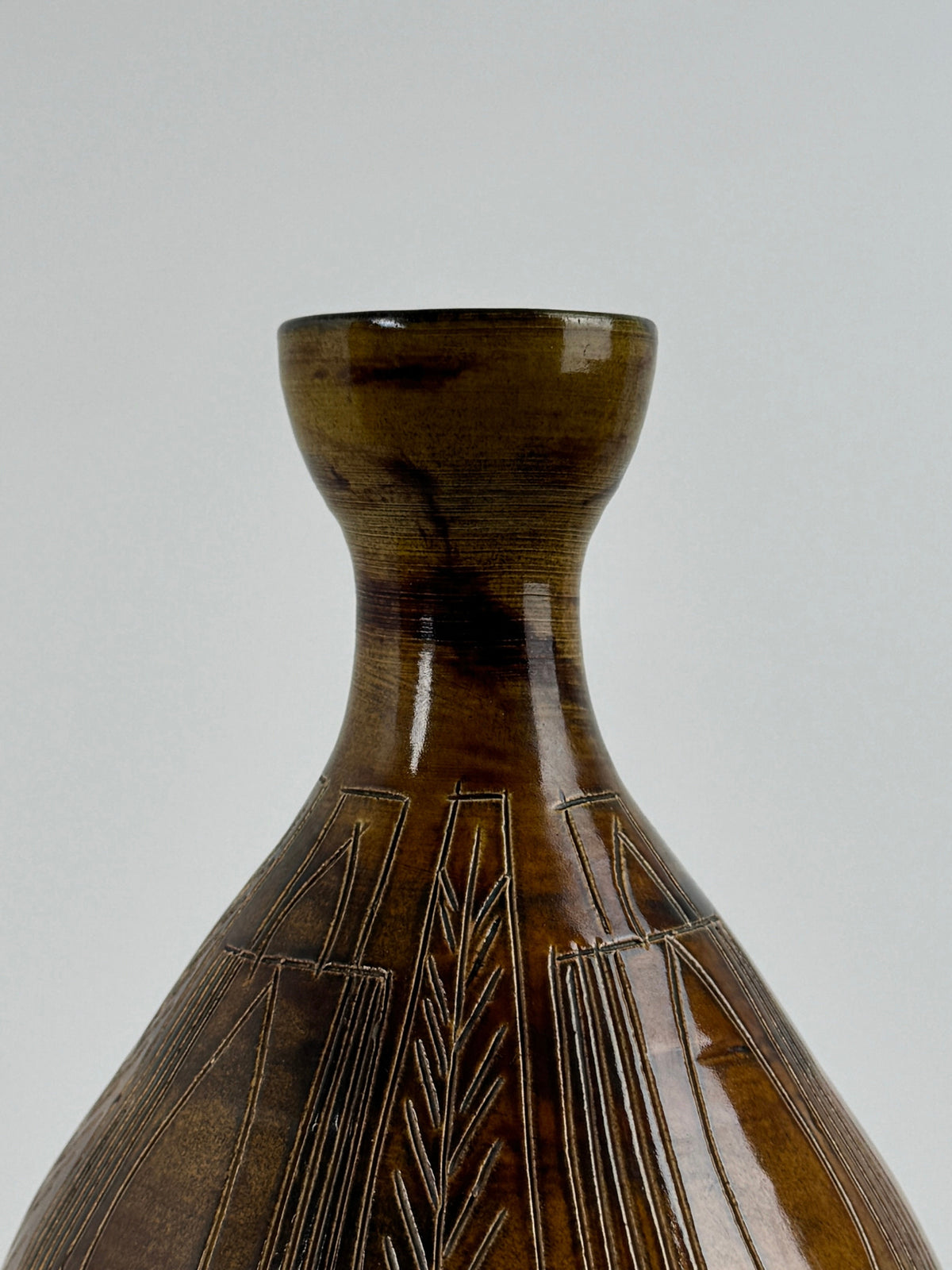 Close-up of a rare Ceramic Vase by the potters of Accolay from Figuresdesign, its glossy brown surface adorned with intricate incised geometrical decoration. The vase features a narrow neck and a flared opening, set against a plain light gray background.