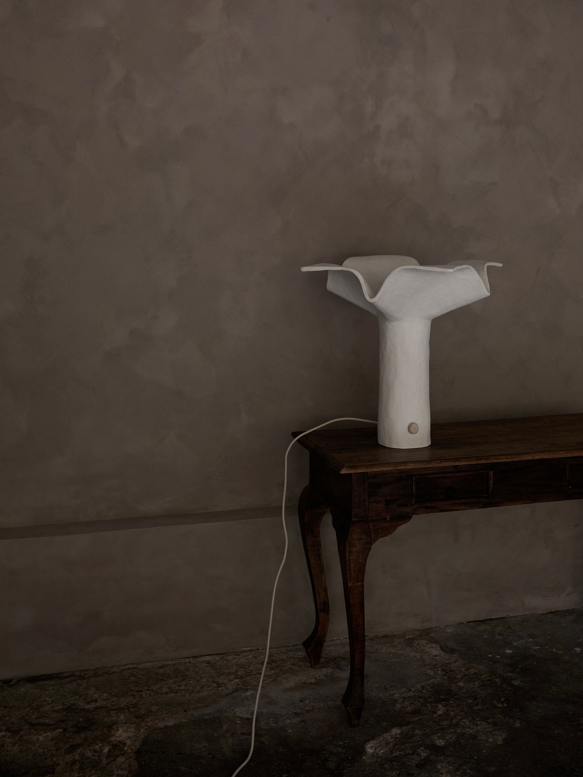 The 'La Fleur' Table Lamp by Studio Tristan Kallas, featuring a distinct white ceramic design, rests elegantly on a dark wood table against a textured gray wall. Its long cord complements the minimalist rustic aesthetic, and floral silhouettes enhance its charm.