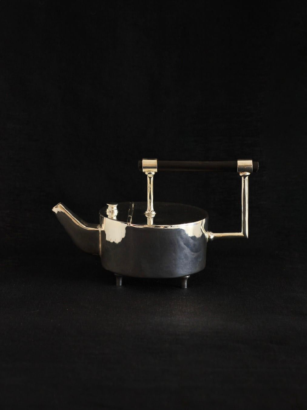 A sleek, modern teapot, named Christopher Dresser's Tea Pot by Boga Avante Shop, features a short and wide silver-plated body. It has a straight spout, an ebonized wood handle, and stands on four small legs. Pictured against a black background, the teapot exhibits a minimalist design with a glossy finish reminiscent of Dr. Christopher Dresser's work.