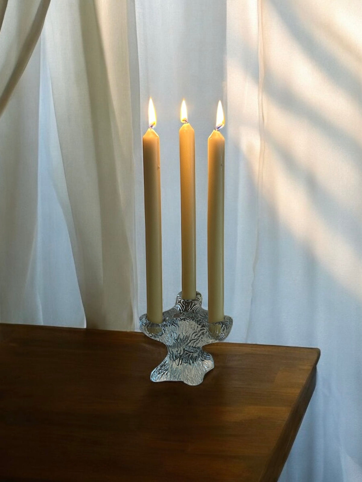 A Sculpted Glass Candelabra by Flock Studio holds three glowing yellow candles on a wooden table. Soft sunlight filters through sheer white curtains in the background, casting gentle shadows.