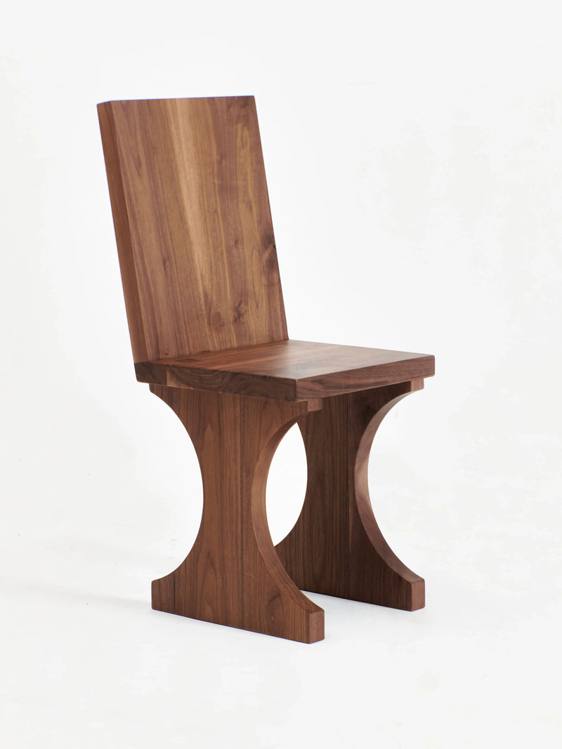 The Portugal Chair No 3 by Project 213A is a modern wooden chair featuring a squared backrest and seat. It stands out with its unique curved supports connecting the seat to the base, which also serve as the legs. Handcrafted from solid walnut, this chair boasts a rich, natural finish against a plain white background.