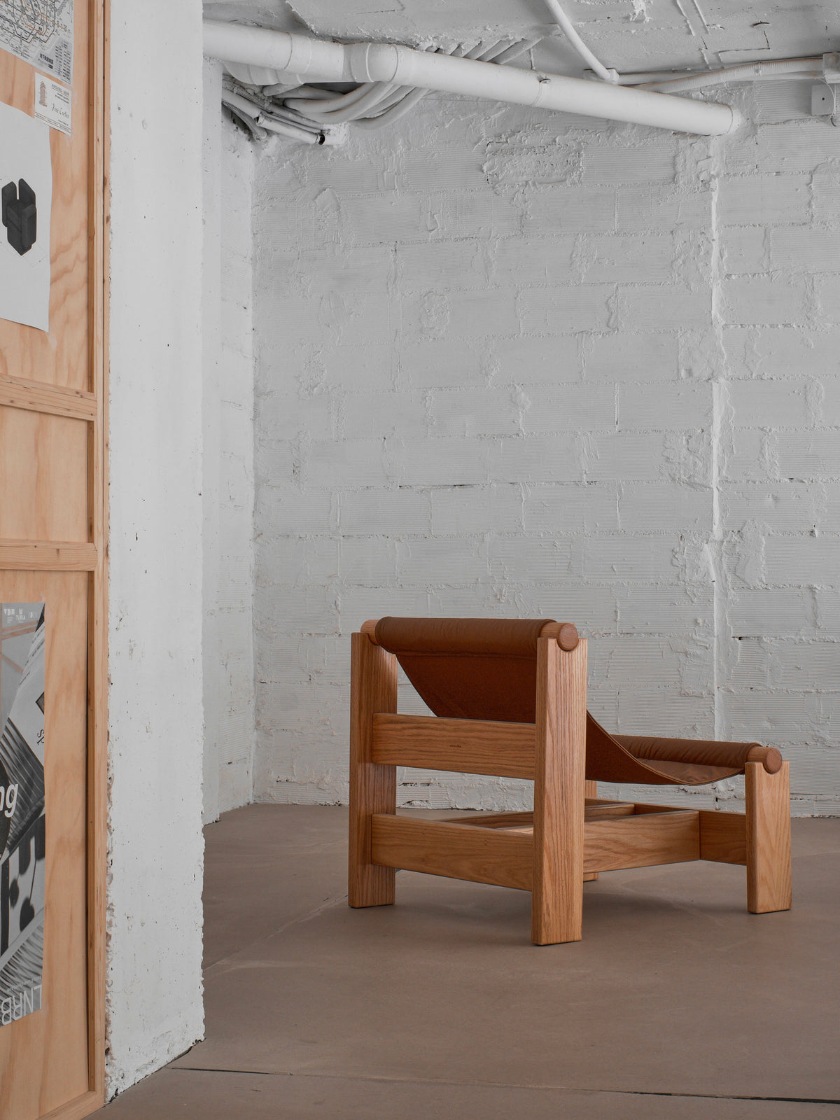 The "Lounge Up" Lounge Chair by malva features a modern, minimalist wood design and brown leather seat for adaptive comfort, ideal for an industrial-style room with white brick walls and exposed pipes.