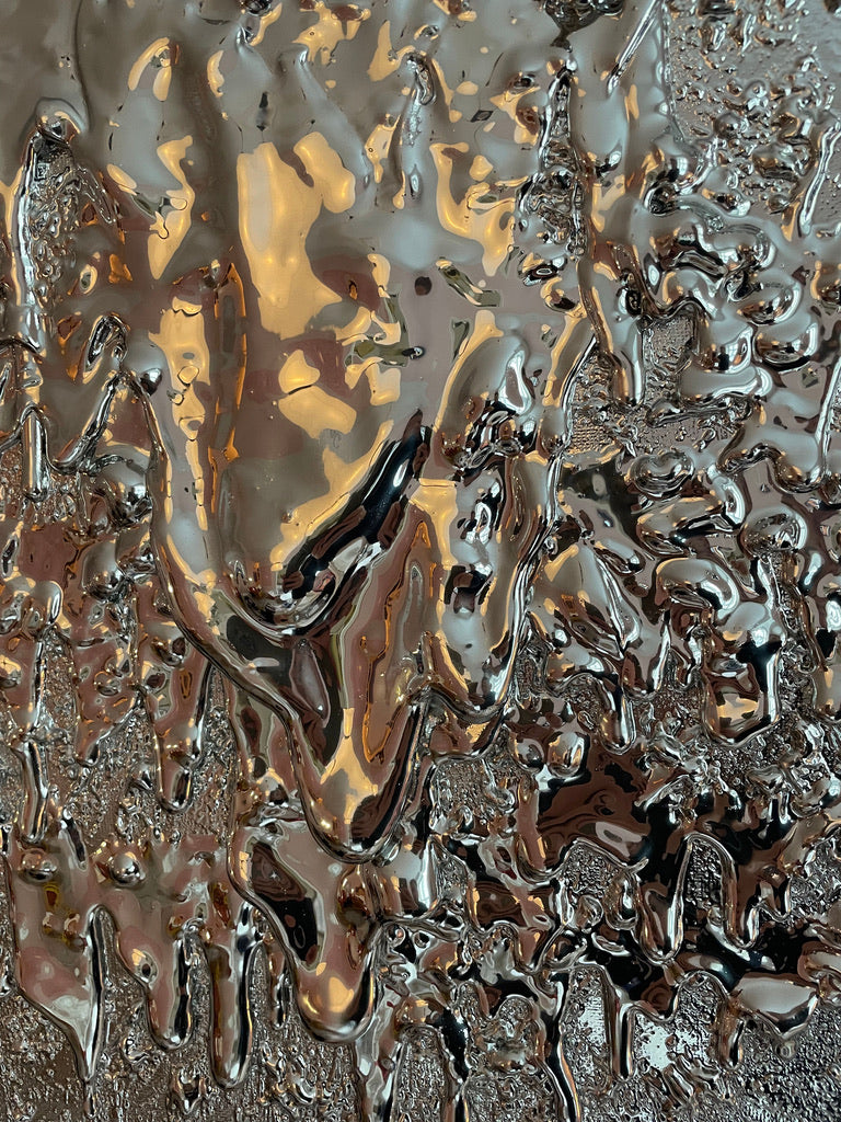 Close-up of John Nordenstein's "Almost a Mirror" sculpture, featuring metallic, reflective surfaces with a fluid, melted appearance. Warm amber and silver hues dominate the piece, creating an organic, textured pattern that resembles both liquid metal and dripped paint—a unique artwork showcasing intricate organic shapes.