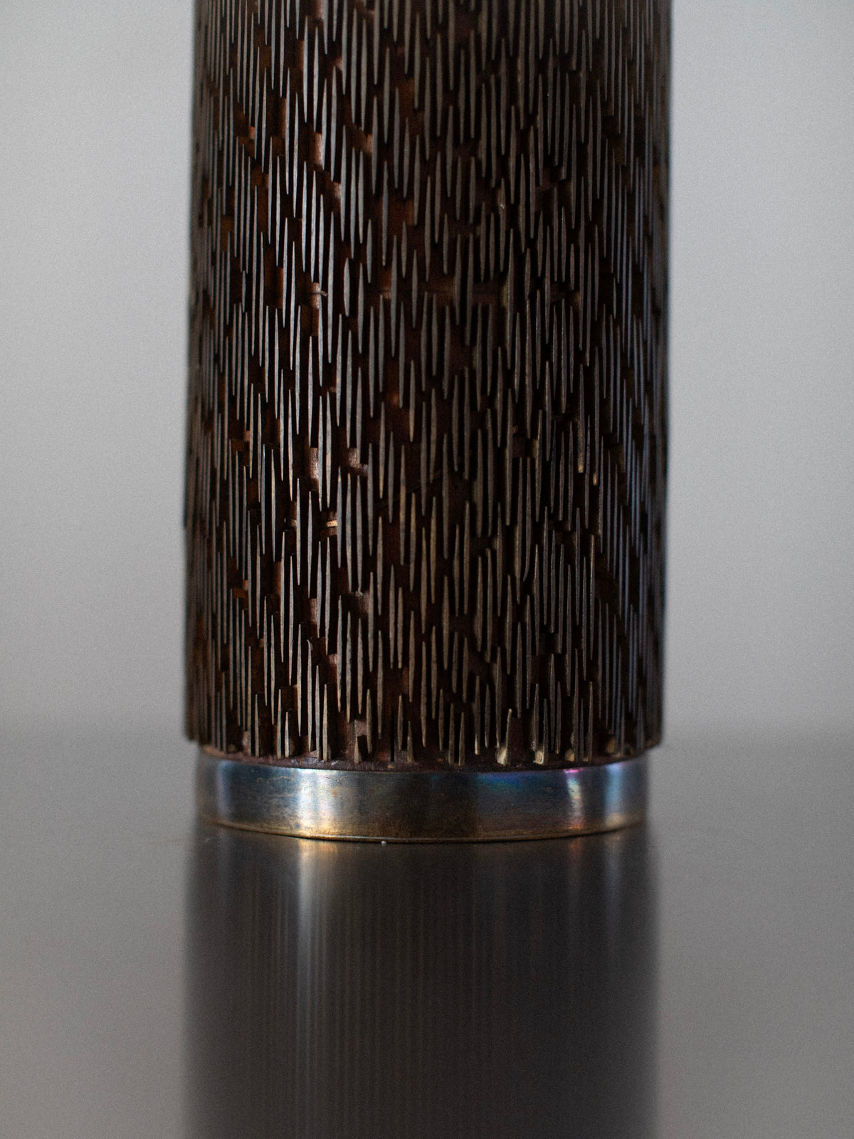 Close-up of the Out For Lunch's Brutalist Floor Lamp, showcasing its textured, bronze metal surface adorned with a pattern of short, vertical lines. The lamp features a smooth, circular metallic base at the bottom. The blurred background highlights the floor lamp’s striking design.