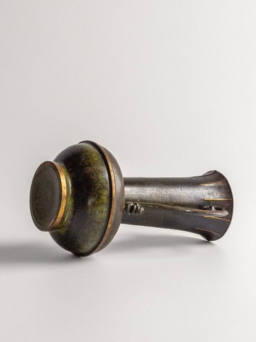 A metal object, possibly an ancient tool or artifact, with a cylindrical handle and a rounded, disk-like end. The object has a dark, weathered patina with greenish and brown hues, resembling the finish of the "Art Deco Bronze Vase" from Spigel's 1930s Scandinavian collection, resting on a plain surface.