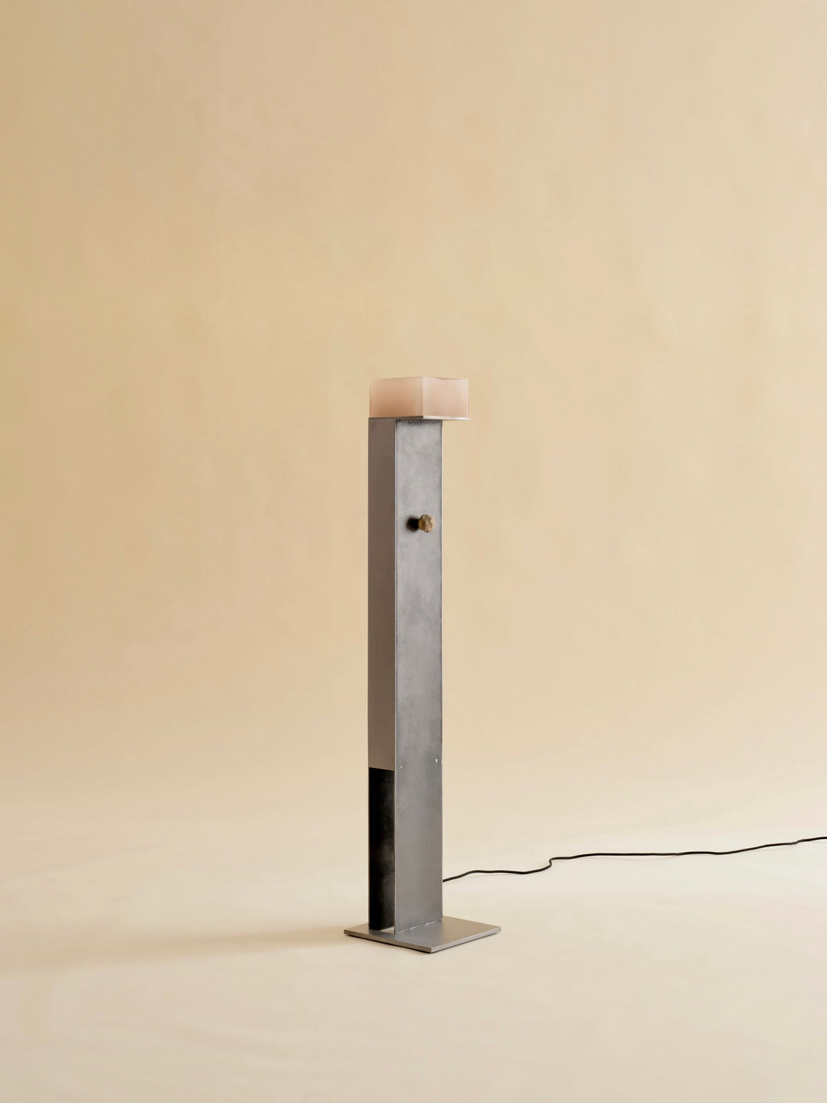 The Perceptions Floor Lamp by Known Work stands on a plain background with a handcrafted rectangular metal base. Its slim column leads to a soft pink, rounded top, illustrating an elegant design with a power cord extending from the base.