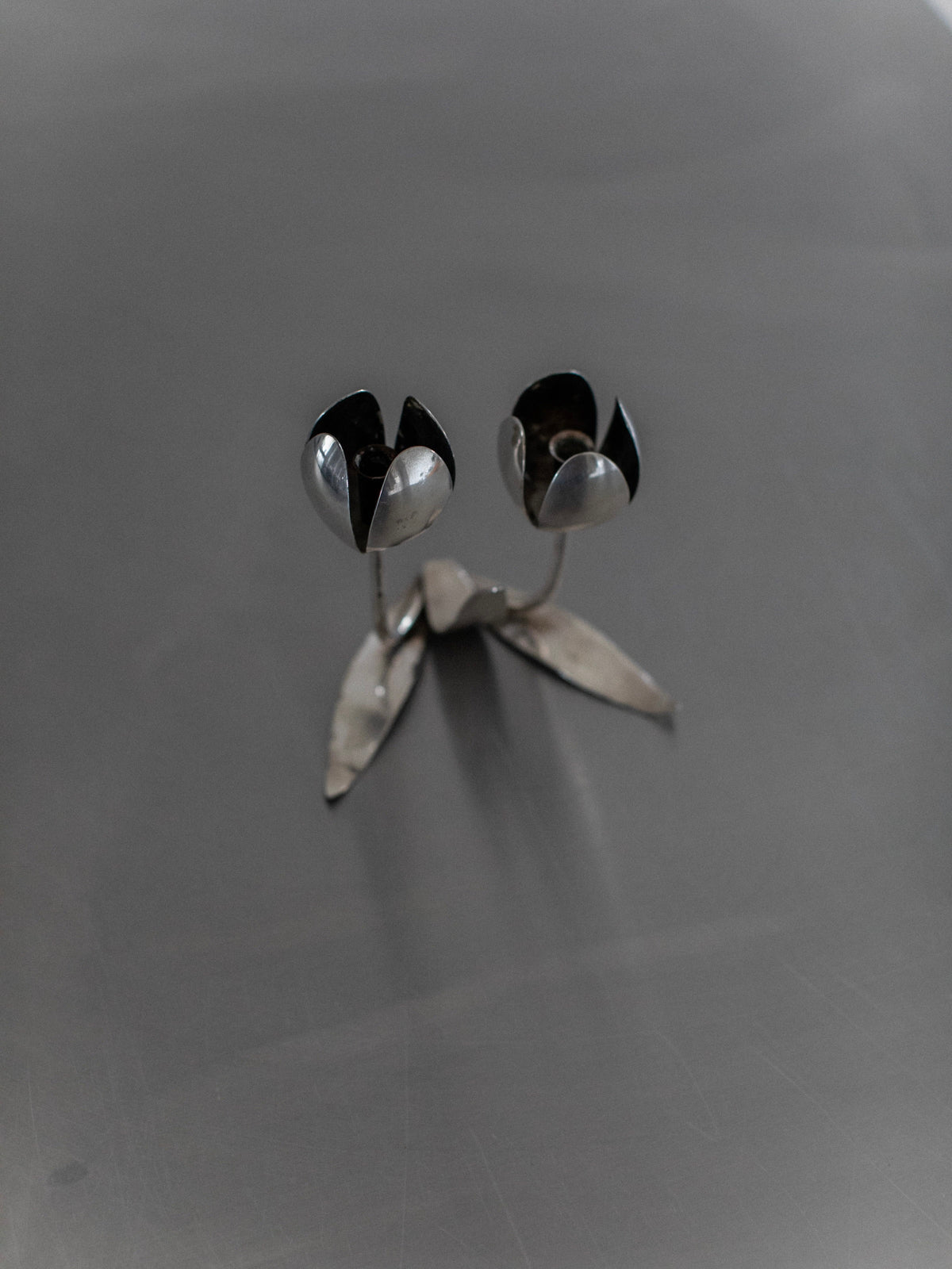 Introducing the Steel Tulip Candleholder by Out For Lunch: This one-of-a-kind, artistic steel piece captures the essence of two abstract flowers with petal-like shapes and a sleek metallic finish. It rests elegantly on a gray base, making it an ideal addition to any modern home decor.