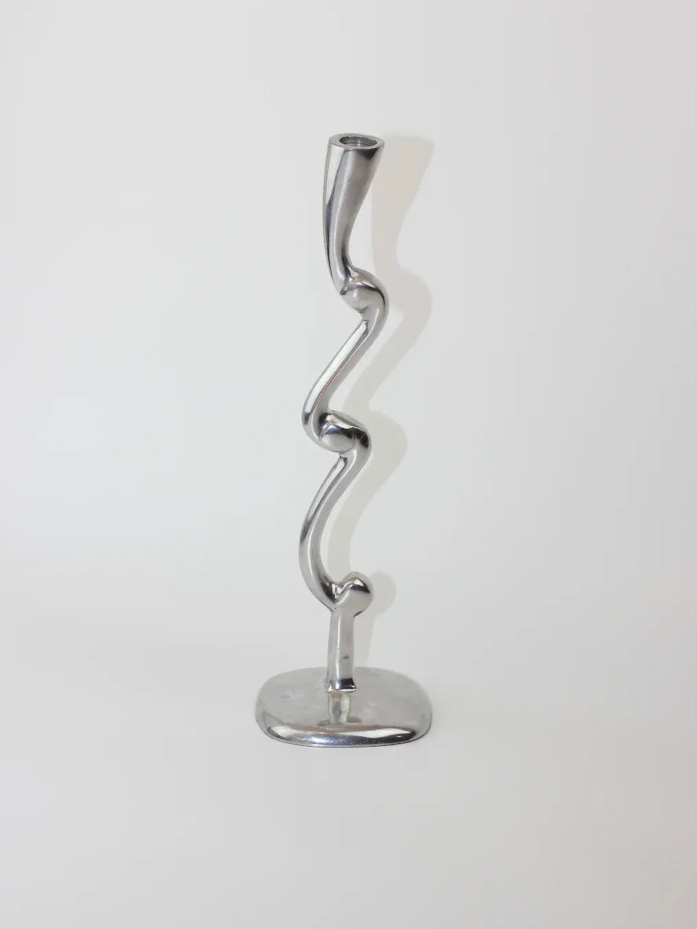 A sleek, silver-hued aluminum candleholder pair from Boga Avante Shop showcases a wavy, sculptural design on a flat base against a simple white backdrop.