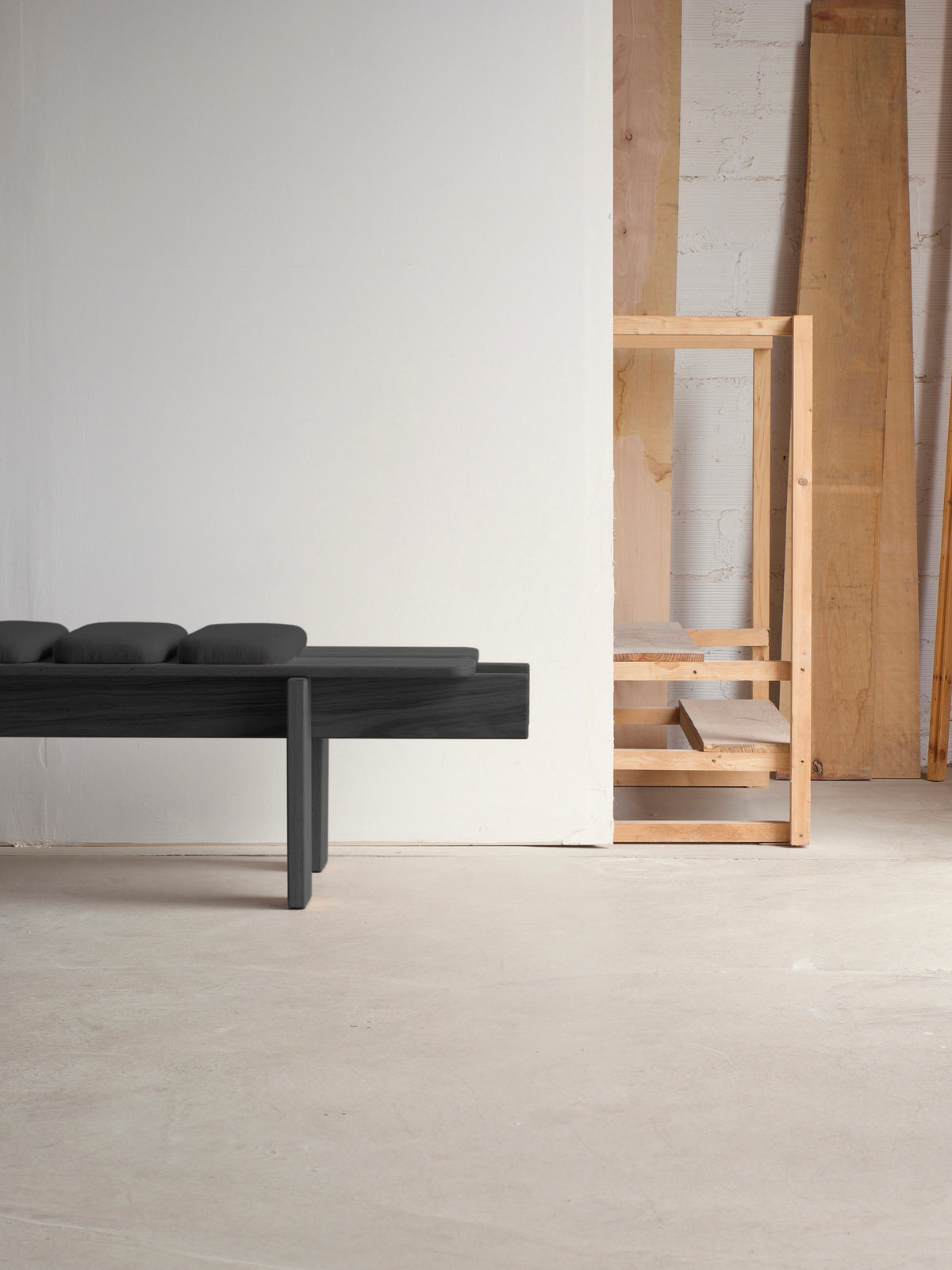 A minimalist interior boasts the Ueno Bench Black Edition by malva on the left with a sleek design and dark cushions. On the right, solid oak boards and a small shelf exemplify Japanese craftsmanship against a light-colored wall, resting on a smooth, pale floor.