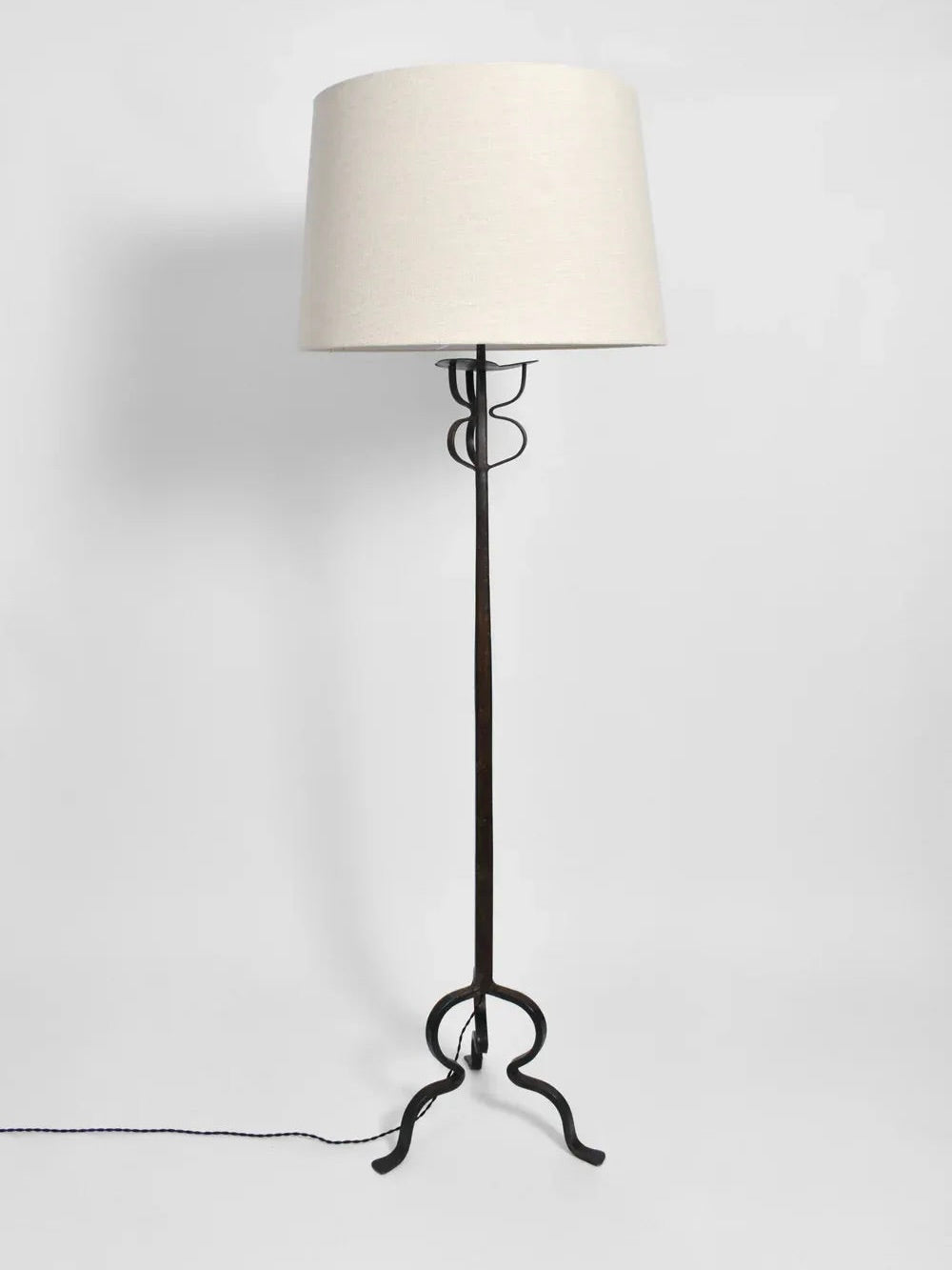 The Tripod Iron Floor Lamp, a creation of Barracuda Interiors reminiscent of 1960s France, boasts a tall black wrought iron frame with a beige cylindrical shade. Its decorative curved base and simple cord add an elegant touch as it stands gracefully against a plain white background.