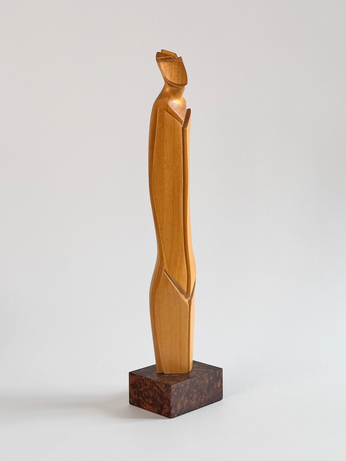 A distinct Light Wood Sculpture by Pierre Dedieu from Figuresdesign features smooth, flowing curves. This minimalist piece stands upright on a dark base and showcases a humanoid form with a small head and elongated body, all set against a light gray background.