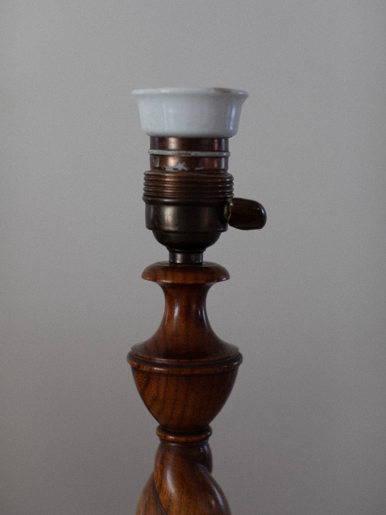 The Early 1900's Table Lamp by Out For Lunch features a wooden base with a metal and porcelain light socket, set against a plain grey background. Missing a bulb or lampshade, the lamp highlights an Art Deco spiral design on its wooden structure, making it ideal for vintage decor enthusiasts.