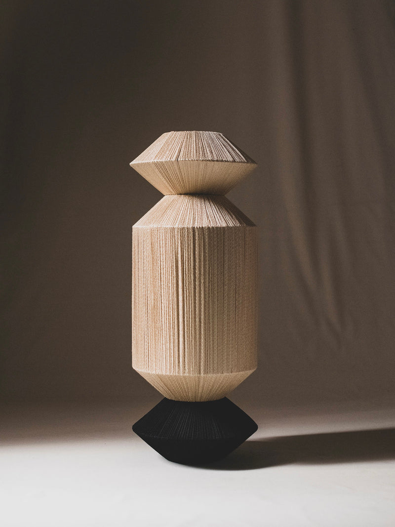 The Column IX Sculpture by ELIURPI features a vertical design made of layered, textured material in natural and dark tones. Its geometric shape, with a central cylinder flanked by two conical forms, is set against a softly lit plain background, ideal for elegant settings.