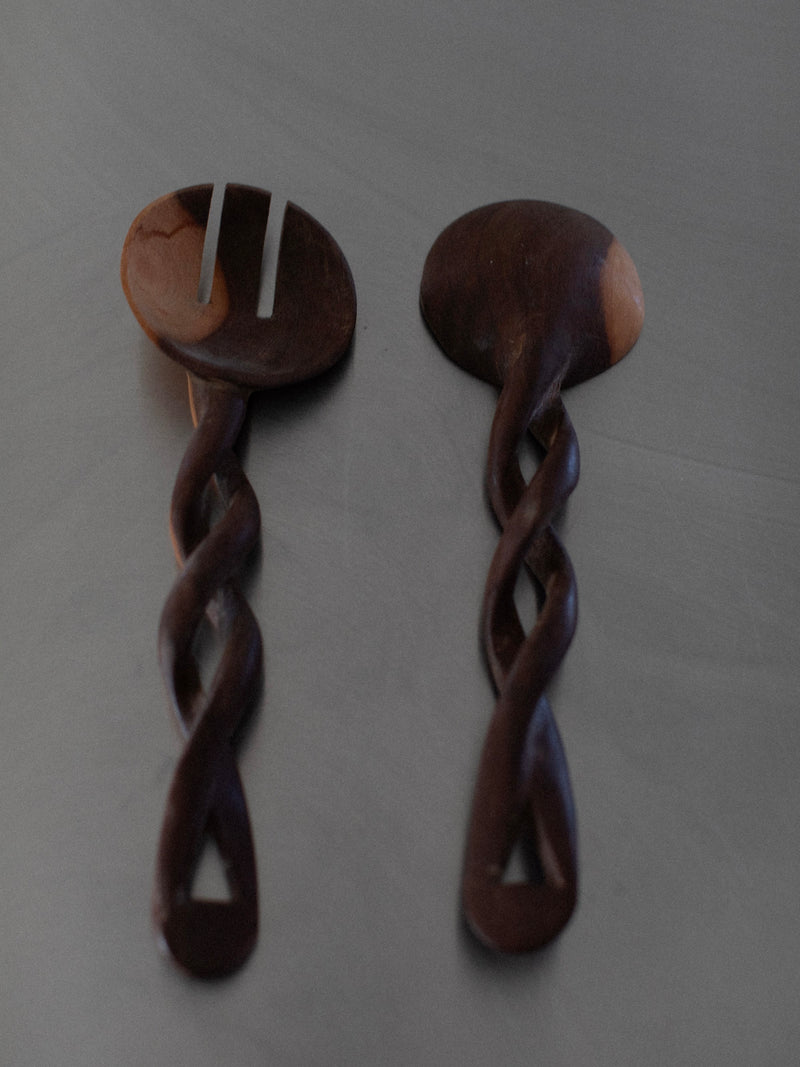 Two handcrafted Twisted Serving Spoons by Out For Lunch with intricate twisted handles rest on a flat surface. One is a serving fork with two prongs, and the other is a serving spoon. The wood appears dark with some lighter patches, showcasing its natural grain and subtle variations from the hand-carving process.