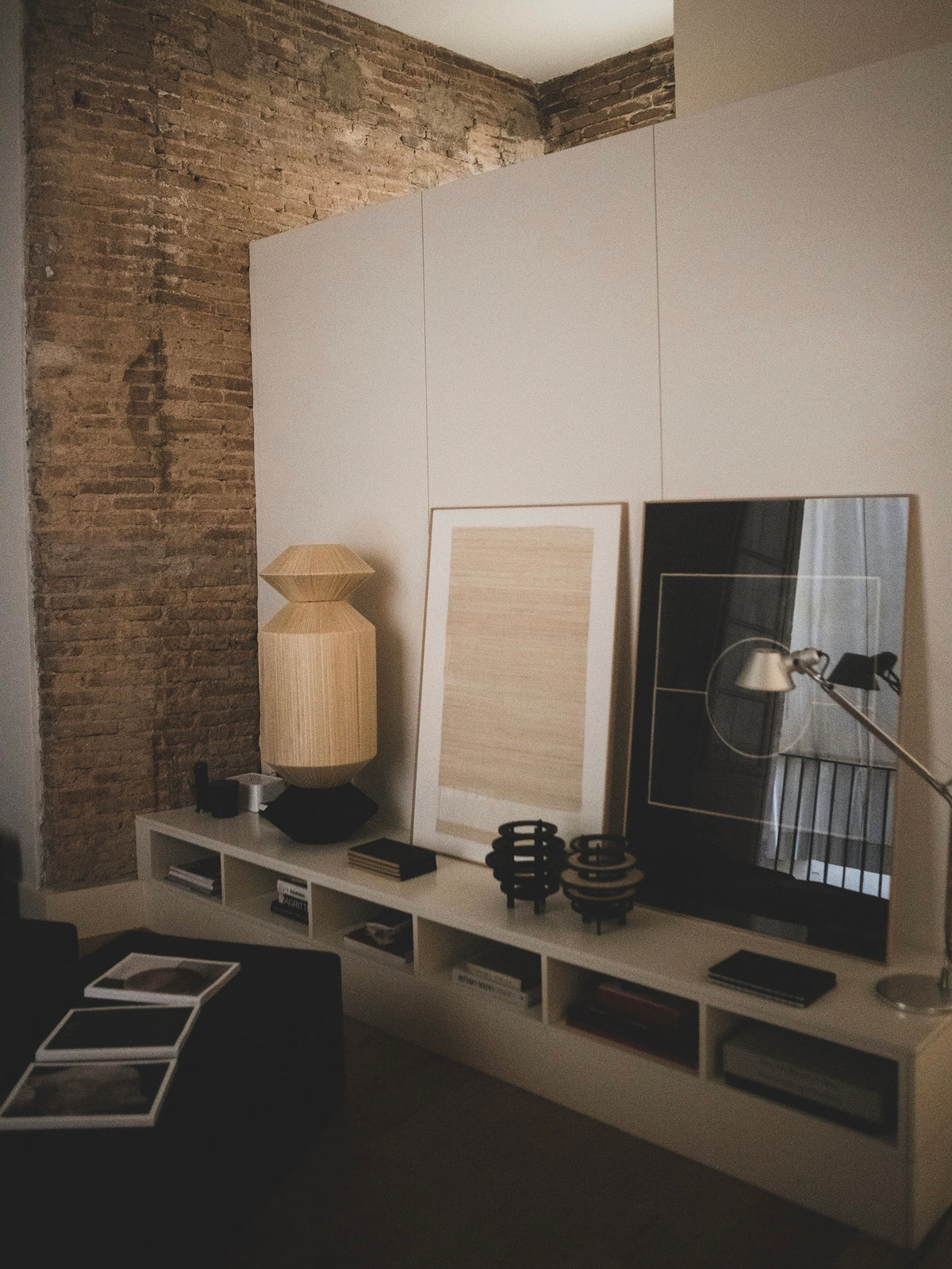 A cozy interior with modern Column IX Sculpture by ELIURPI on a white shelf, set against a brick wall. A tall geometric-designed lamp and a smaller desk lamp enhance the warm and stylish ambiance of this elegant space.
