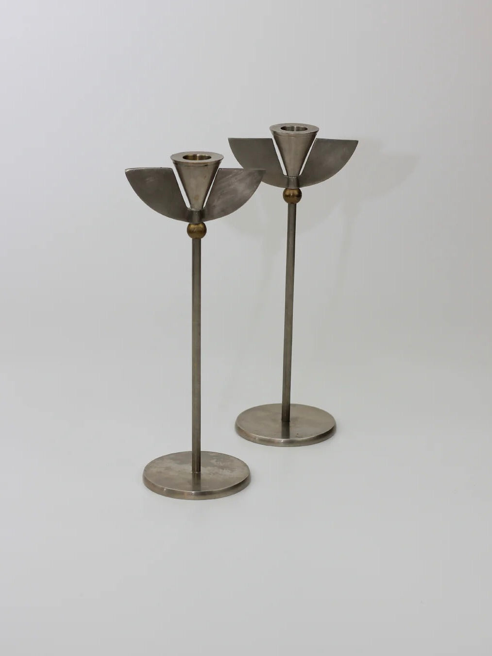 Two modern stainless steel candleholders from Boga Avante Shop, named Memphis Candleholders 80s, feature a geometric design with circular bases, slender stems, and semicircular tops in a Memphis Milano style on a plain white background.
