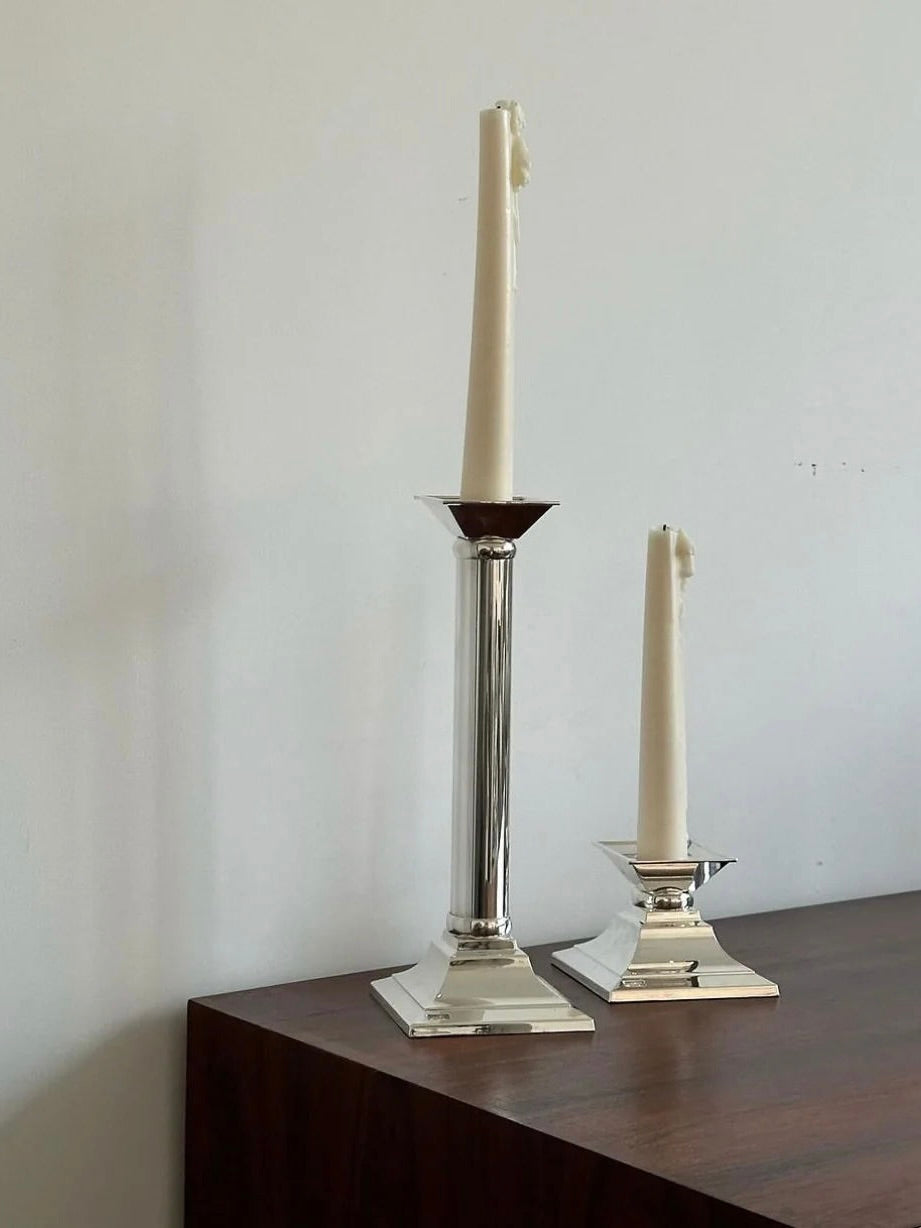 A pair of German Candle Holders by Aesthete Label, designed in a classical column style, hold two white taper candles and are positioned on a dark wooden surface against a plain white wall. One candle holder is significantly taller than the other, creating an asymmetrical visual arrangement. Both candles have slightly melted tops.