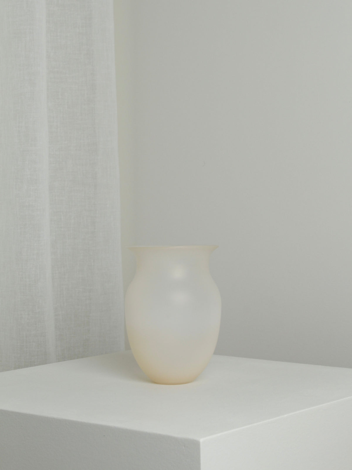 A Medium Vase in Sahara from Verre d'Onge, mouth-blown and translucent white, sits on a white pedestal in a minimalist setting. A white sheer curtain is visible in the background to the left. The overall color scheme is soft and neutral, emphasizing simplicity and elegance in this Sahara-inspired scene.
